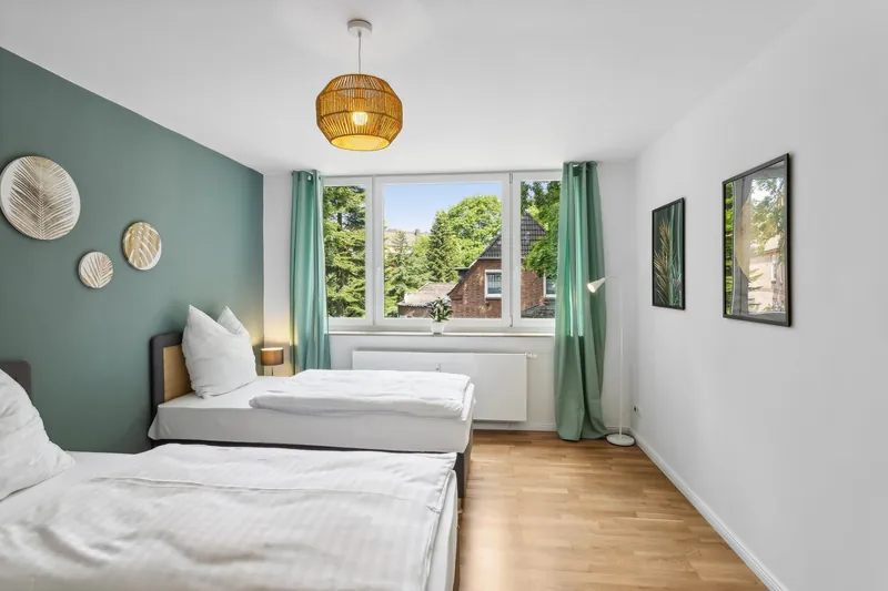 Schlafzimmer mit petrolfarbener Wand, Rattanleuchte und Fenster mit Blick ins Grüne
