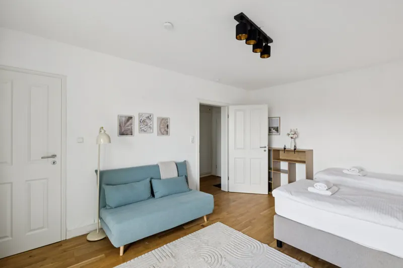 Hellblaues Schlafsofa, daneben ein frisch bezogenes Bett, Stehleuchte und Bilder an der Wand