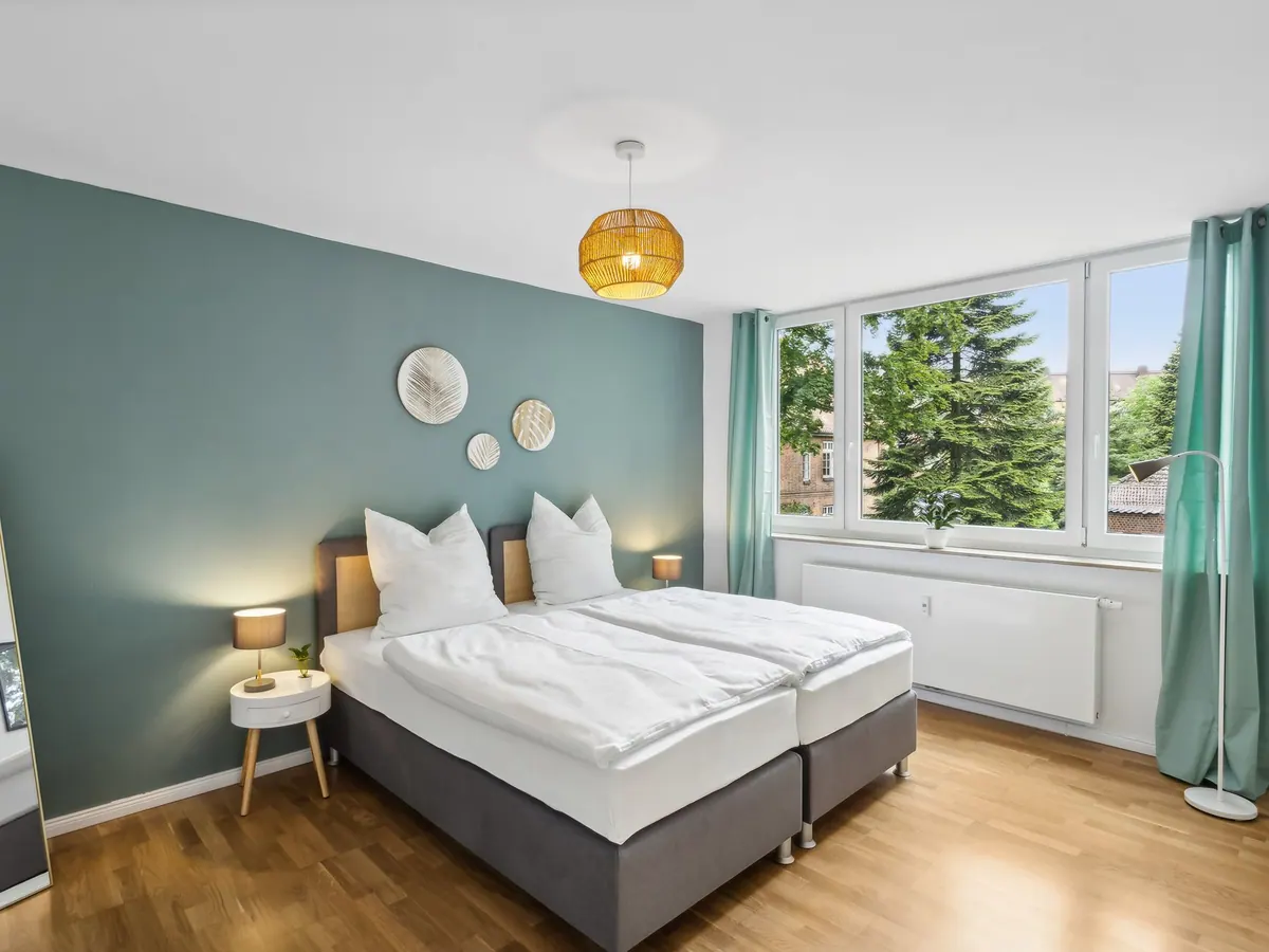 Schlafzimmer mit petrolfarbener Wand, Doppelbett, Rattanleuchte und Fensterfront ins Grüne